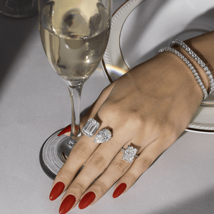Woman wearing 6-carat radiant cut solitaire ring with pavé-set diamond band, styled with champagne glass